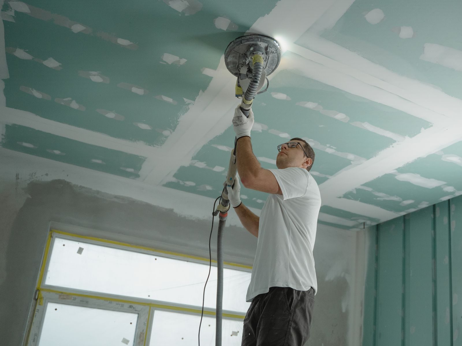 A worker in gloves and safety glasses sanding a drywall ceiling with a pole sander