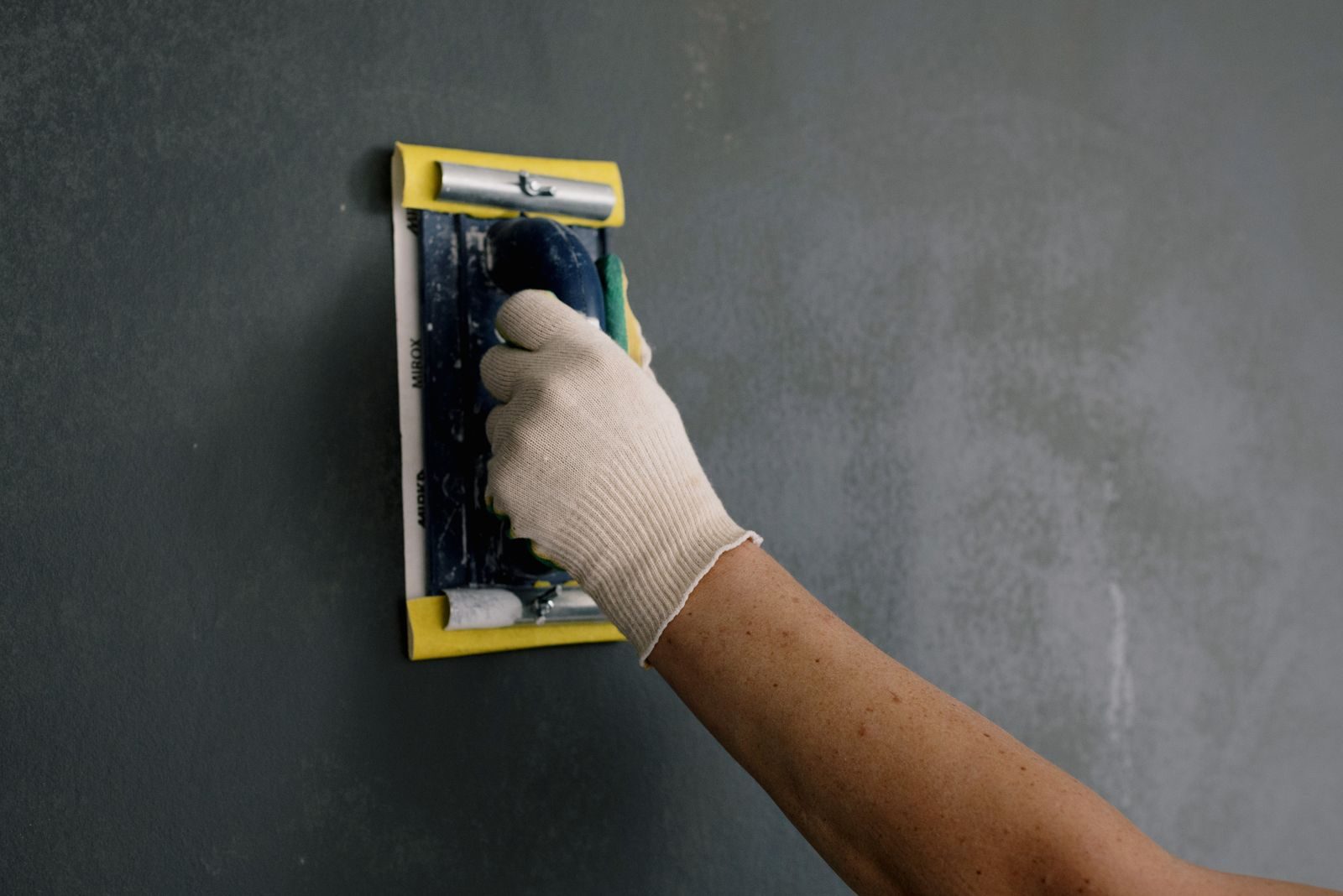 A gloved hand sanding a repaired wall smooth with a sanding block
